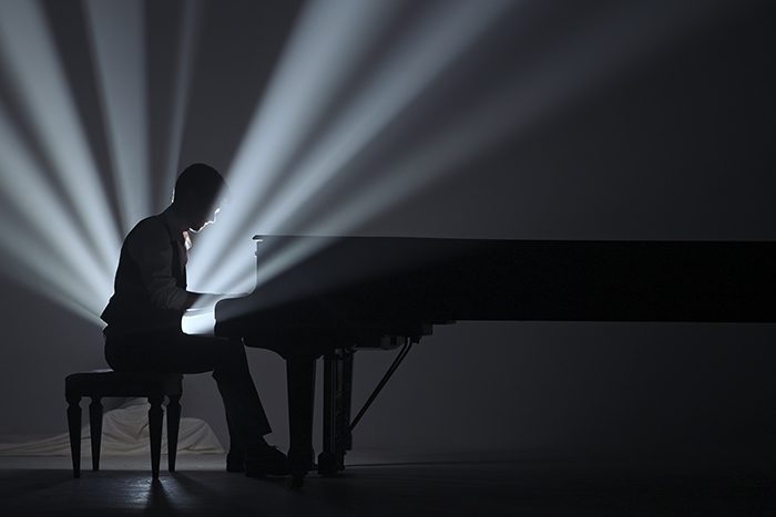 Pianist playing on the black grand piano on dark concert stage against bright spotlight